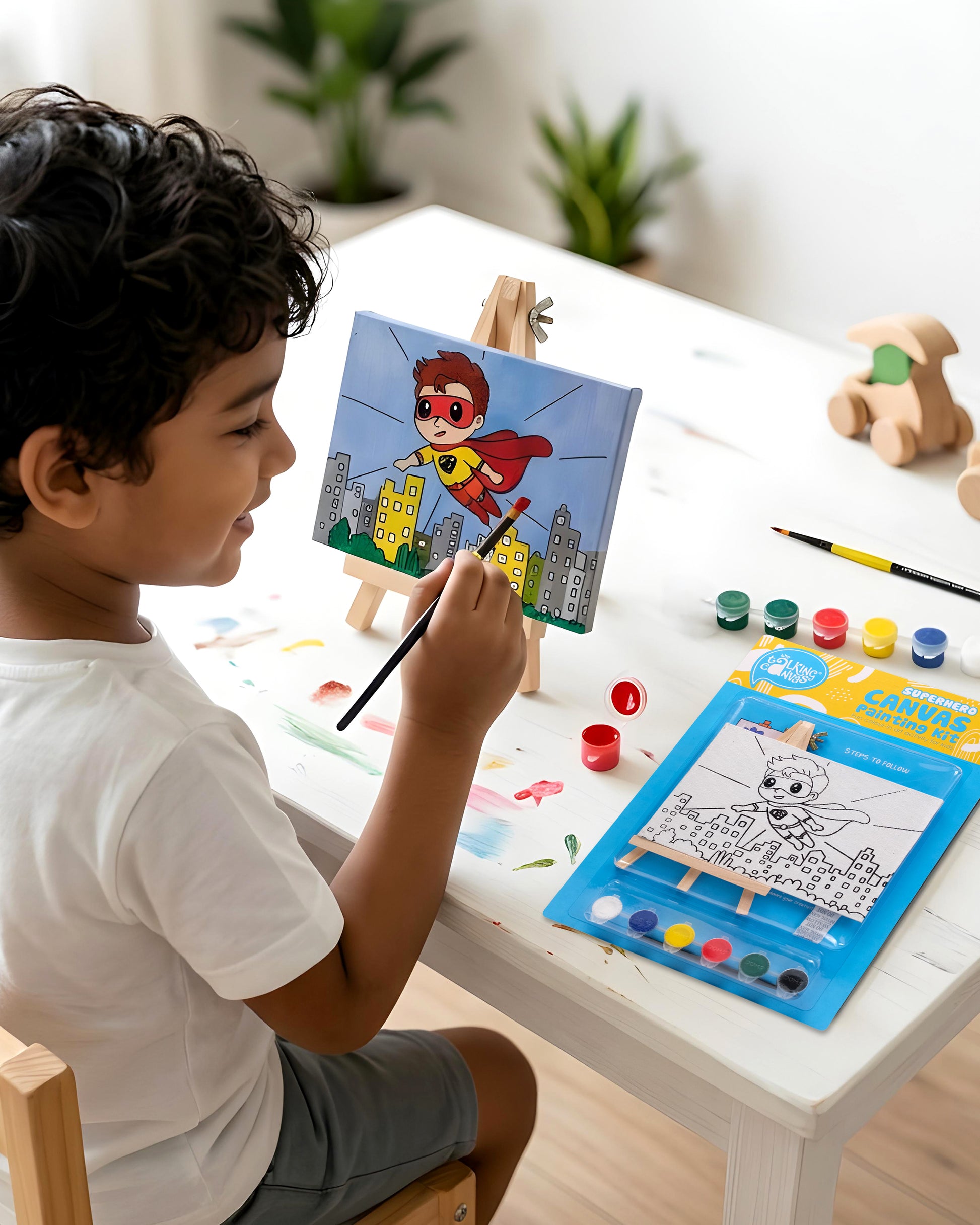 Child painting the Superhero Canvas Painting Kit on a wooden easel using colourful acrylic paints to create a superhero flying above a city skyline during a fun screen-free art activity.